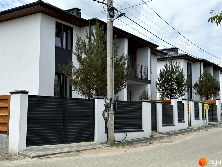 Котеджне містечко VillaTwins: фасади білих будинків з темними вікнами та балконами. Перед будинками огорожа з темних панелей та дерева.