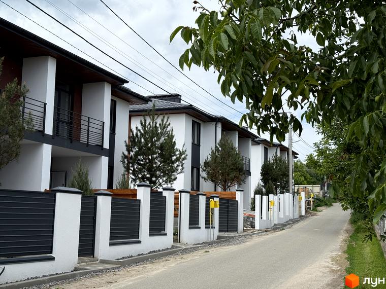 Ряд білих двоповерхових будинків Котеджного містечка VillaTwins з темними дахами та балконами вздовж дороги. Територія облаштована огорожами та зеленими насадженнями.