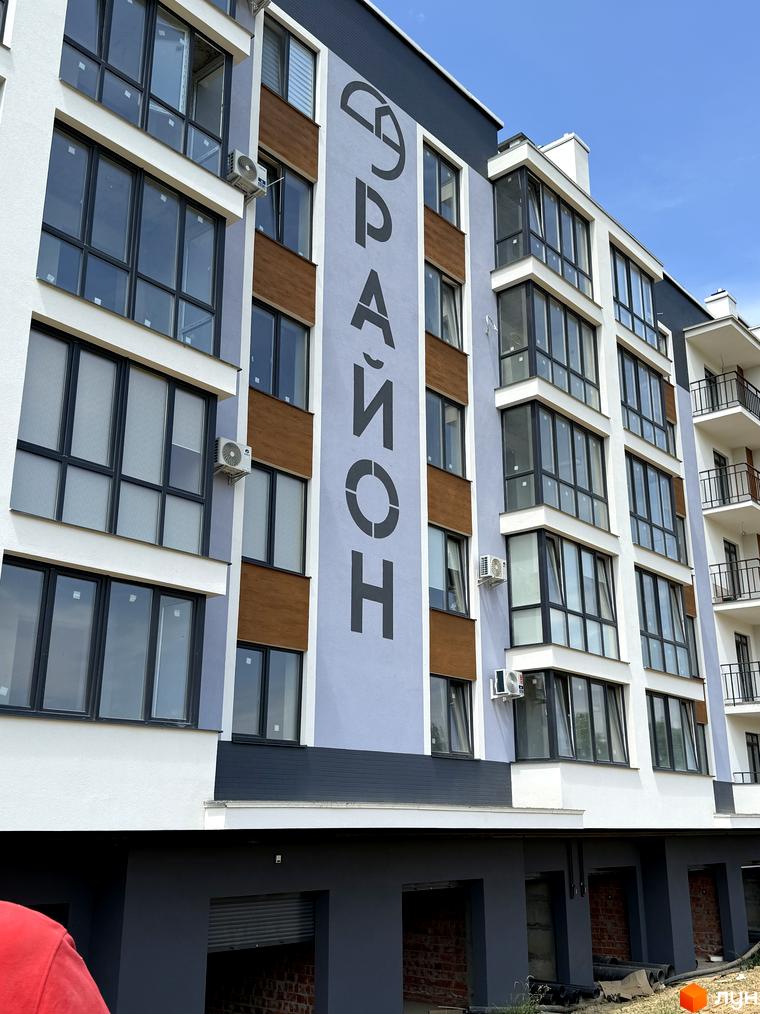 Зовнішній вигляд новобудови ЖК 9 Район з великими вікнами та балконами. Будинок має сіро-білий фасад з дерев'яними вставками.