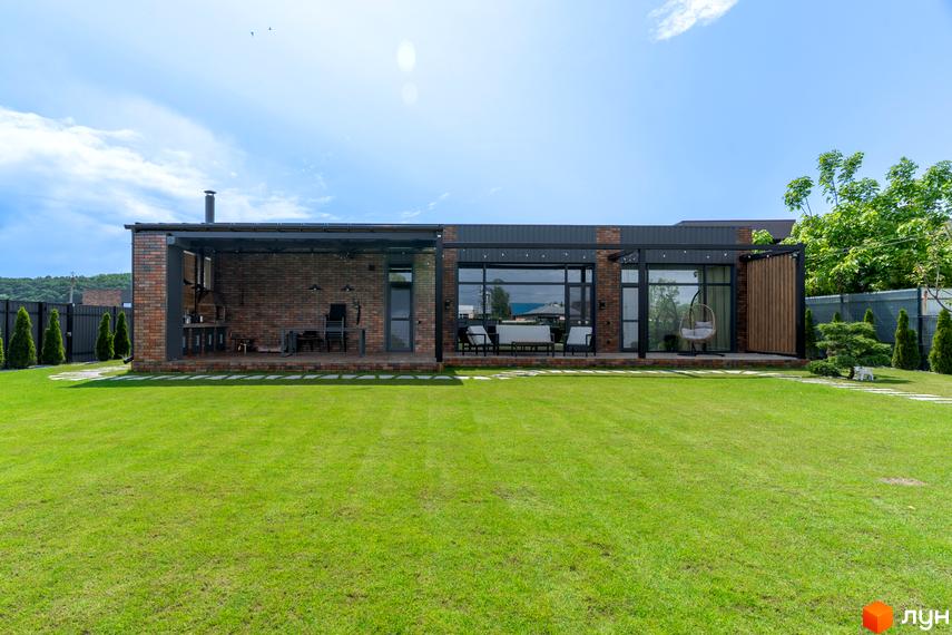 Котеджне містечко КМ Озерний: одноповерховий будинок з цегляним фасадом та великими вікнами, накрита тераса з зоною барбекю та відпочинку, зелений газон перед будинком.