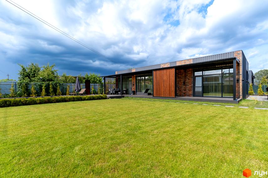 КМ Озерний: одноповерховий будинок з цегляним фасадом та великими вікнами, на тлі зелений газон та дерева. На території розташовані меблі для відпочинку.