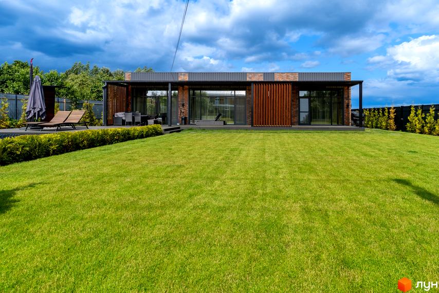 Котеджне містечко КМ Озерний: одноповерховий будинок з великими вікнами та дерев'яними елементами фасаду, поруч зелений газон та зона відпочинку з лежаками.