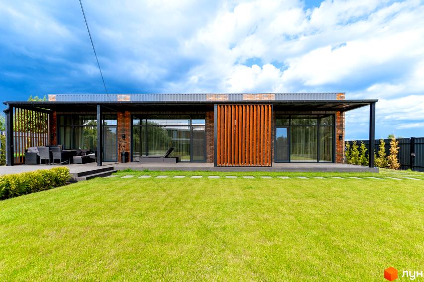 Котеджне містечко Озерний: одноповерховий будинок з цегляними стінами, великими вікнами та терасою. Прибудинкова територія з зеленим газоном та доріжкою з плитки.