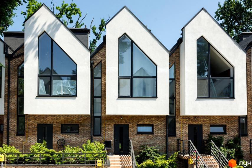 Котеджне містечко КМ Park Residence 3: фасади будинків з великими трикутними вікнами та цегляною кладкою. Зелена прибудинкова територія з огорожею.