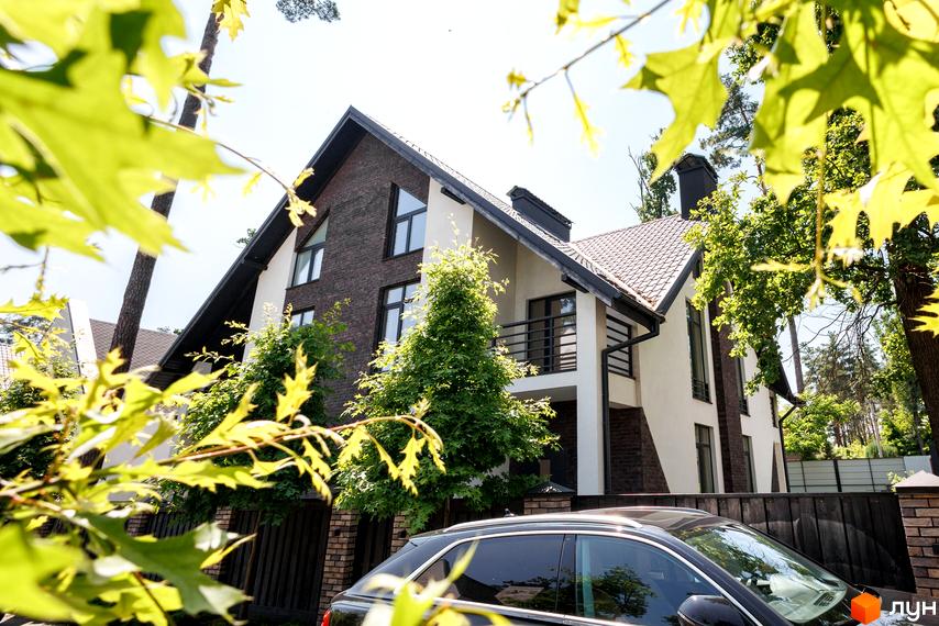 Котеджне містечко КМ Park Residence 3. Фасад будинку з цегляними та білими елементами, балкон, вікна, зелені дерева та паркан.