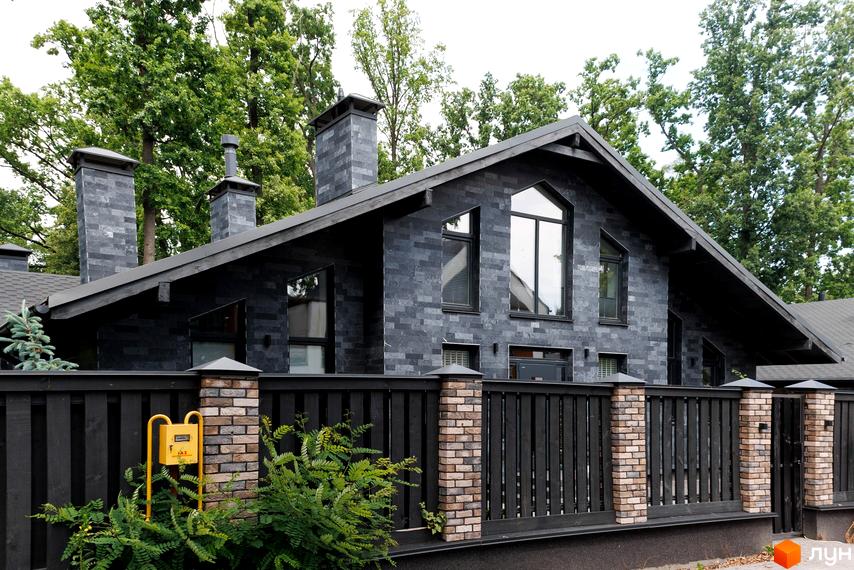 Котеджне містечко КМ Park Residence 3. Фасад будинку облицьований темною плиткою, великі вікна та димарі. Огороджена територія з парканом та зеленими насадженнями.