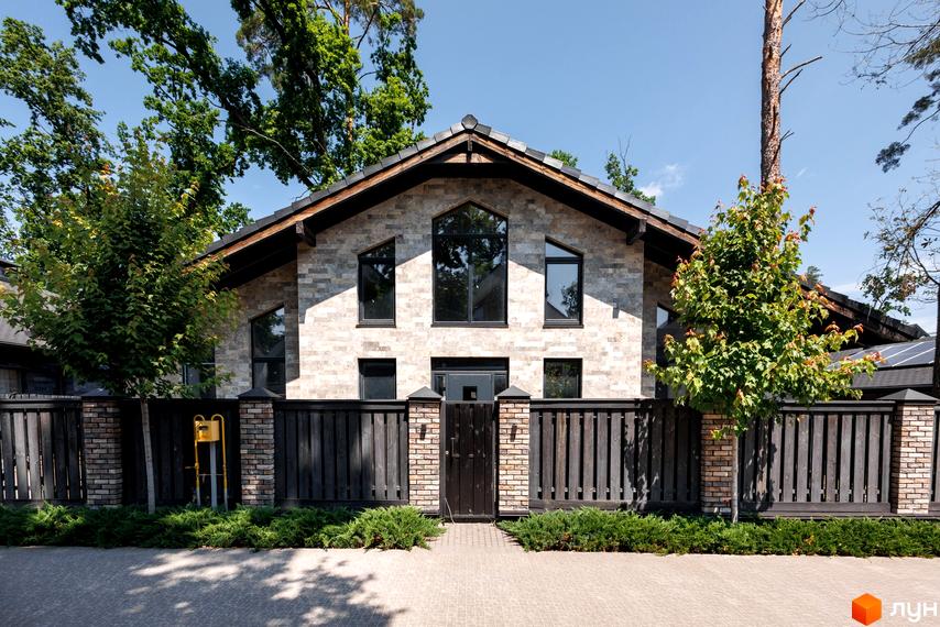 Котеджне містечко КМ Park Residence 3. Фасад будинку з сірої цегли з великими вікнами та темним парканом навколо. Навколо будинку зелені дерева та кущі.