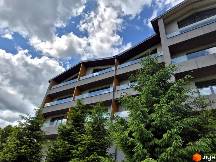 Апарт-комплекс EverGreen з сірими фасадами, скляними балконами та зеленими ялинами на передньому плані. Сучасна архітектура новобудови під блакитним небом з хмарами.