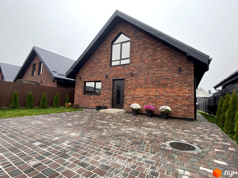 Цегляний будинок з чорним дахом та вікнами. Перед будинком викладена бруківка, є клумби з квітами.