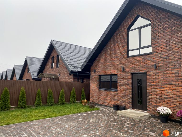 Зображення сторінки житлового комплексу КМ Баварія Village. Цегляні будинки з чорними дахами, великими вікнами та вхідними дверима. Перед будинками доглянута територія з газоном, туями та бруківкою.