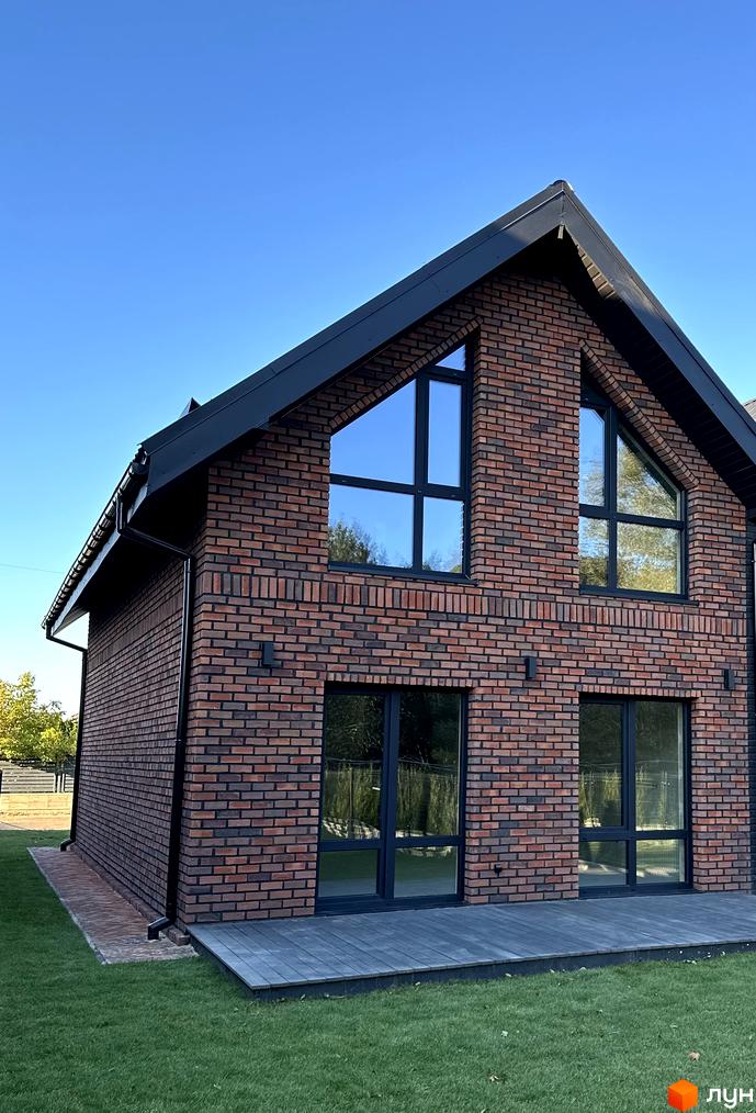 Двоповерховий будинок з цегляними стінами та панорамними вікнами. Поряд з будинком газон та тераса з дерев'яним покриттям.