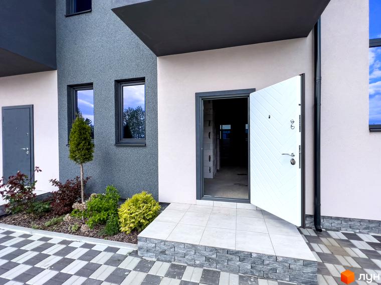 Зовнішній вигляд будинку КМ Одеський з відкритою білою дверима та вхідною групою. Навколо будинку розташовані зелені насадження та доріжка з плитки.
