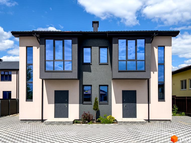 Котеджне містечко КМ Одеський: двоповерхові будинки з темними дахами, великими вікнами та сірими дверима на тлі блакитного неба з хмарами. Перед будинками викладений плиткою двір.