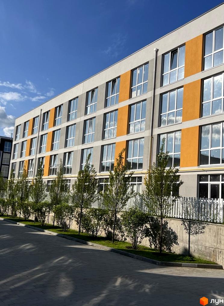 Багатоповерховий будинок ЖК Sanville Park з великими вікнами та помаранчевими вставками на фасаді. Перед будинком доглянута прибудинкова територія з деревами та газоном.