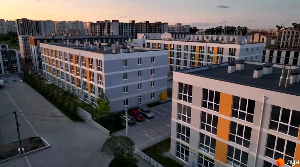 Зовнішній вигляд новобудови ЖК Sanville Park з сірими та жовтими фасадами, вікнами та парковкою. Навколо комплексу зелені насадження та сіра огорожа.