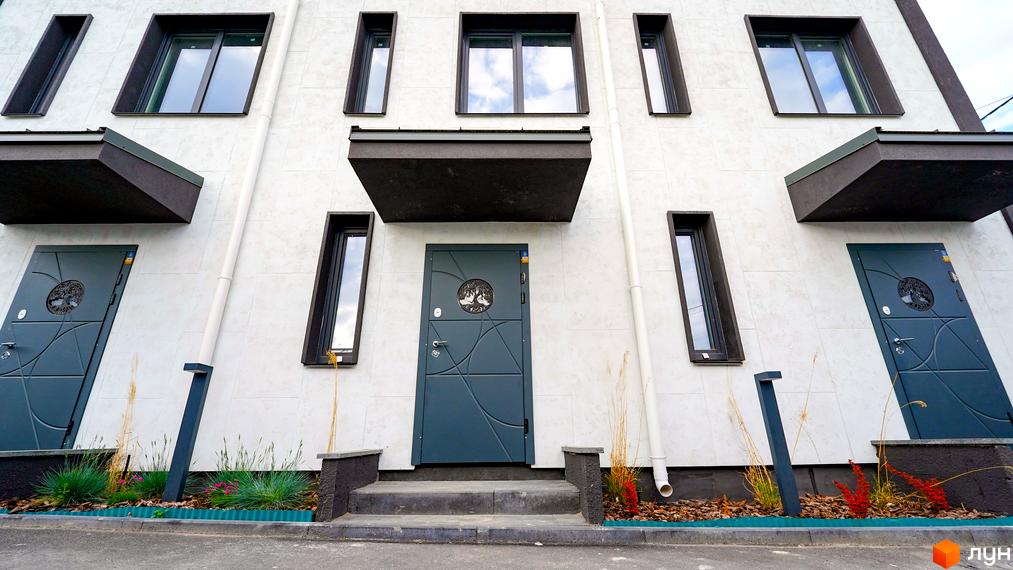 Фасад таунхауса New Smart 15 з сірими дверима та вікнами, оточений зеленню та декоративними рослинами.