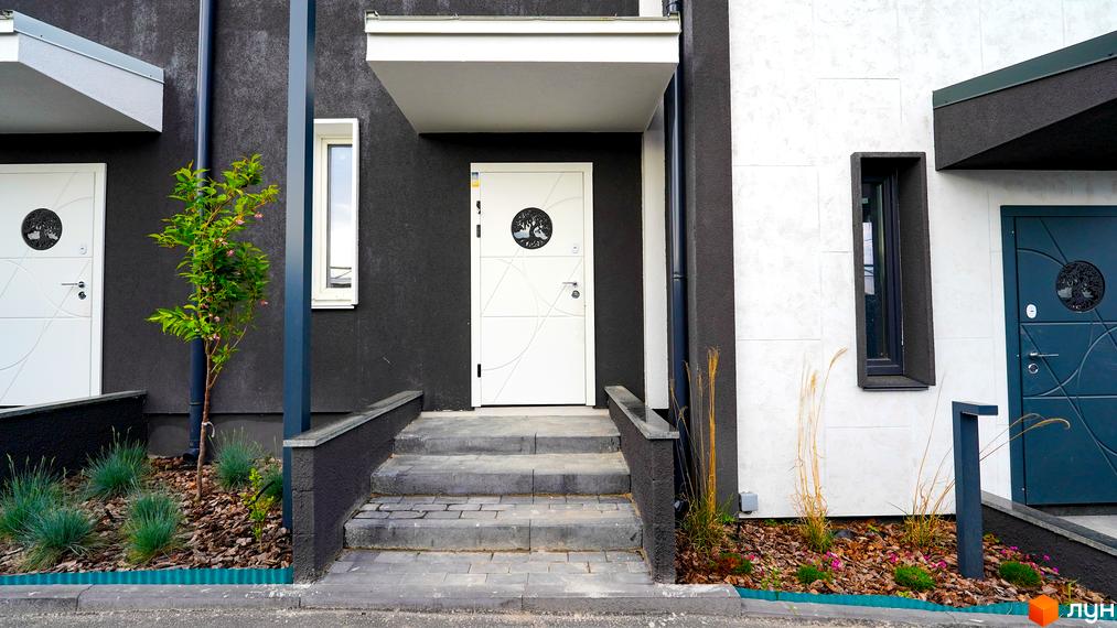 Білий вхід до будинку з чорними стінами, сходами та деревом. На подвір'ї висаджені рослини та квіти, що створюють затишну атмосферу.
