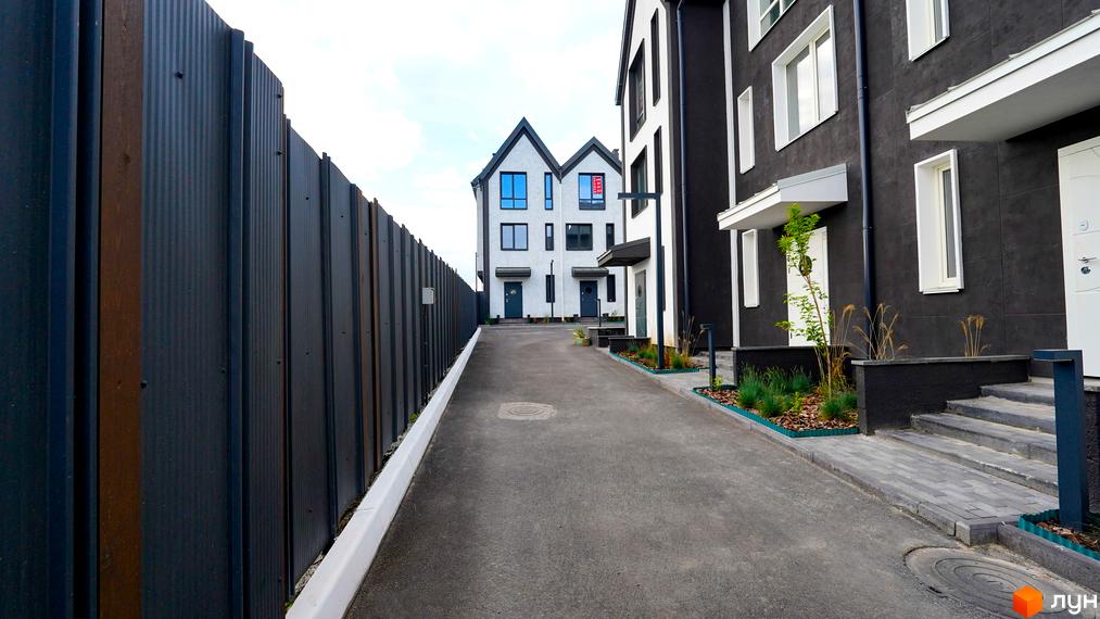 Фасад таунхауса New Smart 15 з темними стінами та білими вікнами. Дорога з асфальтовим покриттям веде до будинків, поруч розташований паркан.