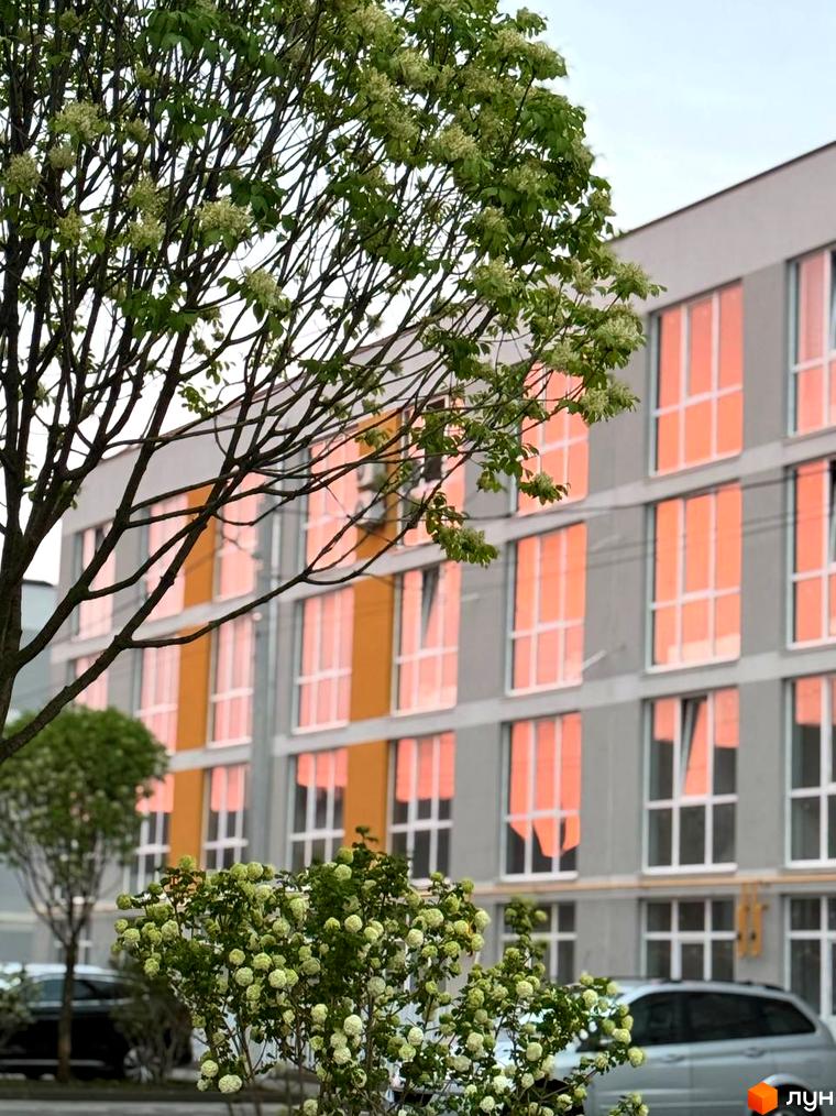 Житловий комплекс Sanville Park з великими вікнами, що відбивають світло. На передньому плані зелені кущі та дерева, що створюють затишну атмосферу.