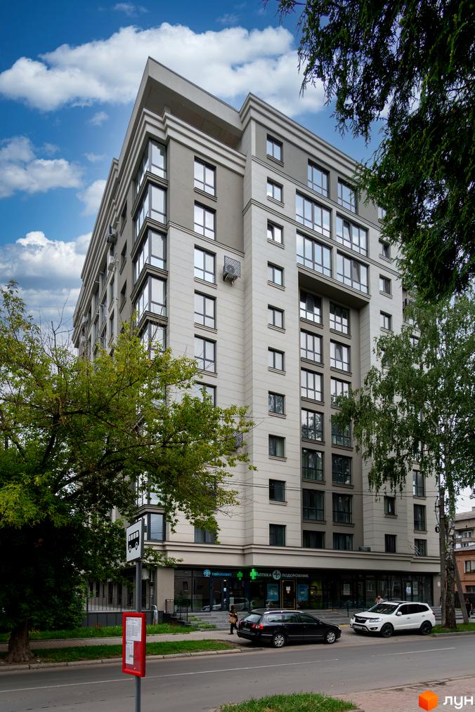 Зовнішній вигляд новобудови ЖК King Hall з великою кількістю вікон та сірим фасадом, зелені дерева та тротуар поруч.