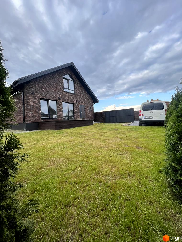 Котеджне містечко КМ Одноповерховий Київ - Віта Поштова. Цегляний будинок з великими вікнами та дерев'яною терасою на фоні зеленого газону та хмарного неба.