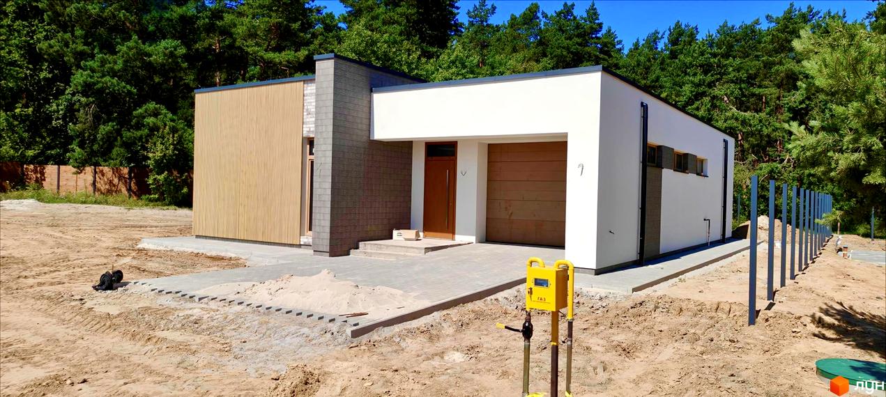 Котеджне містечко LOTUS. Зовнішній вигляд будинку з дерев'яною та цегляною обробкою, гаражними воротами та вхідними дверима. Прибудинкова територія з парканом та газовим лічильником.