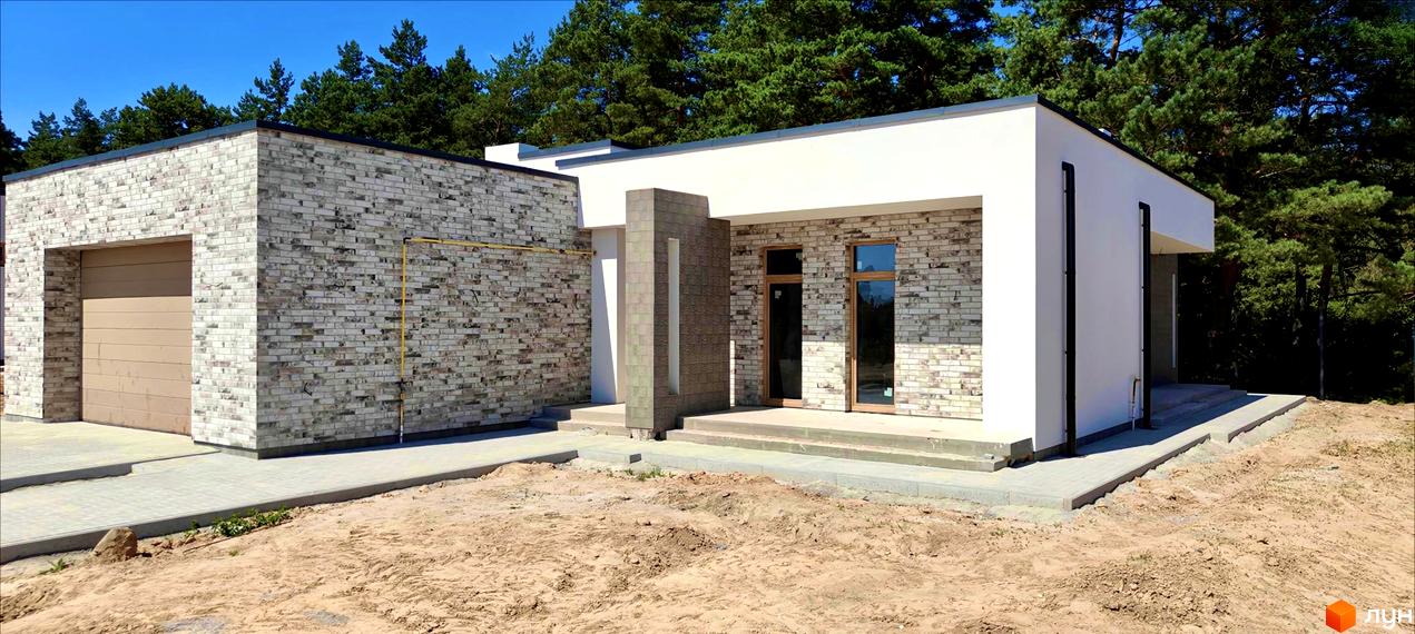 Котеджне містечко LOTUS: будинок з цегляним фасадом та гаражем на тлі соснового лісу. Територія навколо будинку вкрита піском та вимощена плиткою.