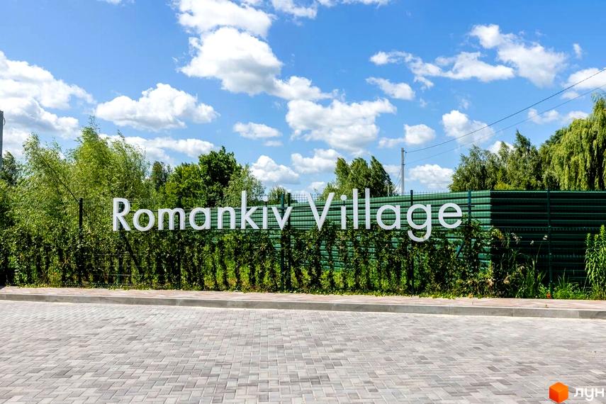 Котеджне містечко Romankiv Village. Білі об'ємні літери назви комплексу на фоні зеленого паркану та зелені. Перед комплексом вимощена сіра бруківка.