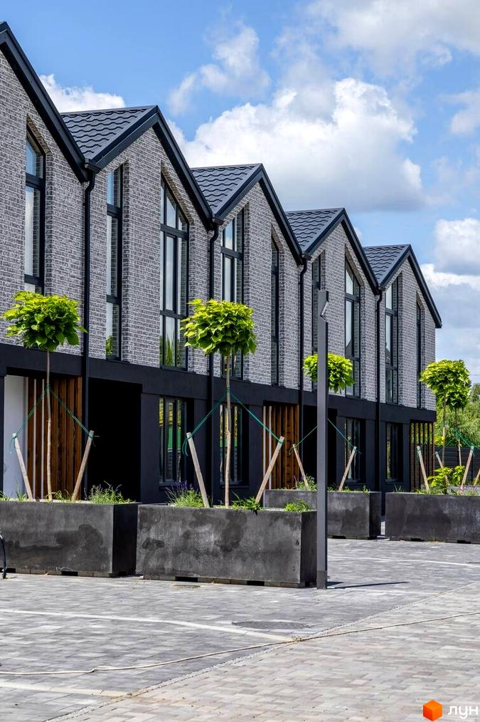 Котеджне містечко КМ Romankiv Village з сірої цегли під сірим дахом та облаштованою прибудинковою територією з деревами у великих бетонних вазонах.