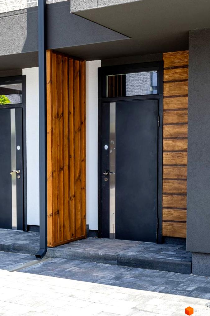 Зовнішній вигляд входу в котеджне містечко КМ Romankiv Village з темними дверима та дерев'яною обшивкою. Територія перед будинком вимощена плиткою.