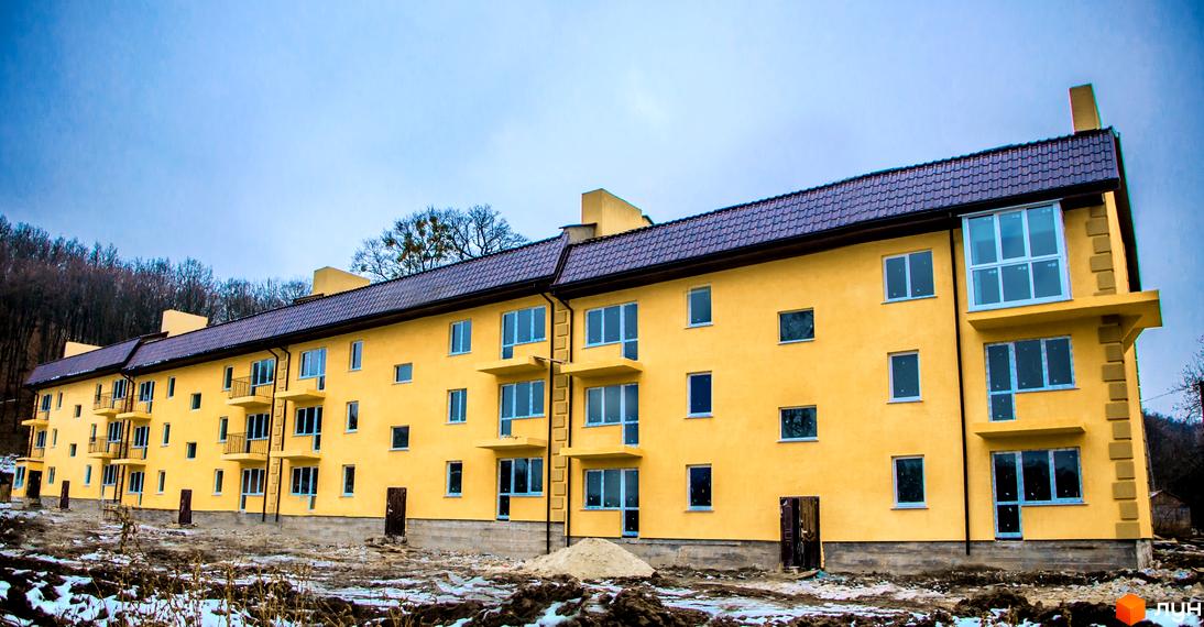 Фасад жовтого багатоквартирного будинку з коричневим дахом та балконами. Територія навколо будинку вкрита снігом та землею.