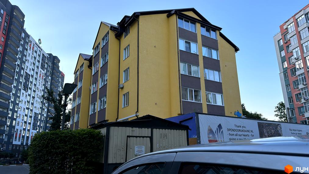 Фасад житлового комплексу Академквартал-3 у жовтих та сірих тонах, поруч розташовані багатоповерхові будинки, видно частину прибудинкової території з деревами та автомобілем.