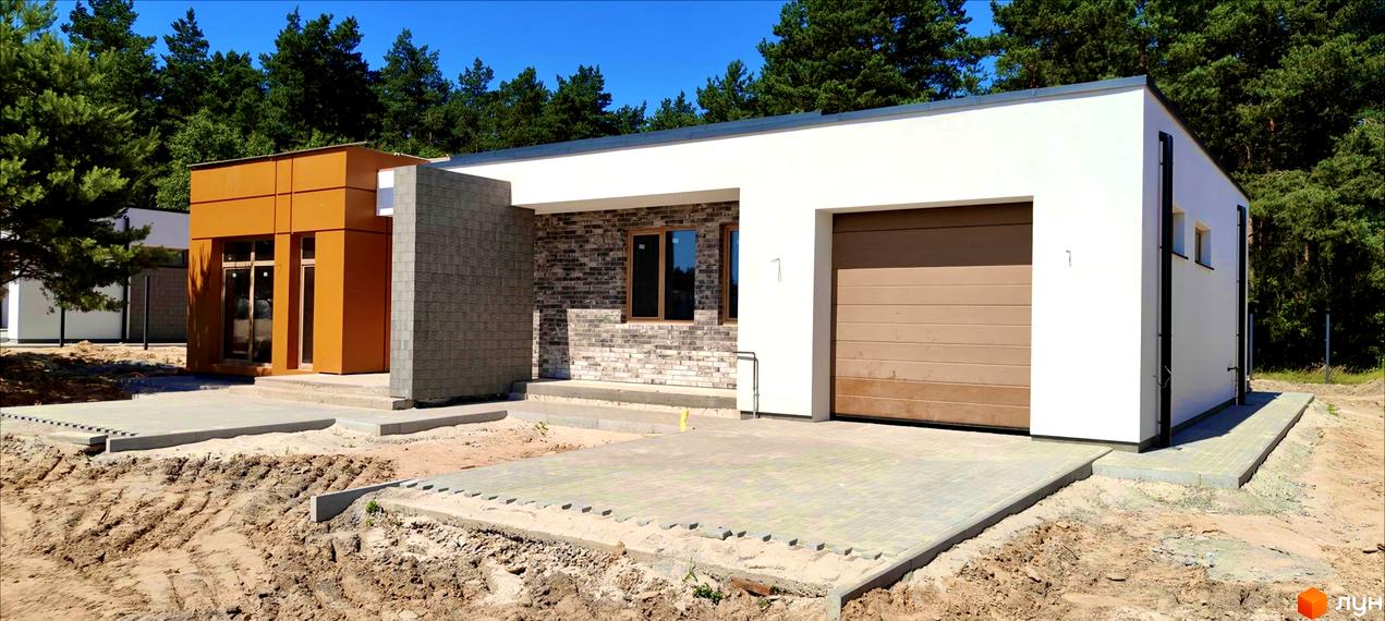 Зовнішній вигляд будинку KM LOTUS з цегляними та білими фасадами, оранжевим блоком та гаражними воротами. Навколо будинку облаштована прибудинкова територія з бруківкою та зеленими насадженнями.