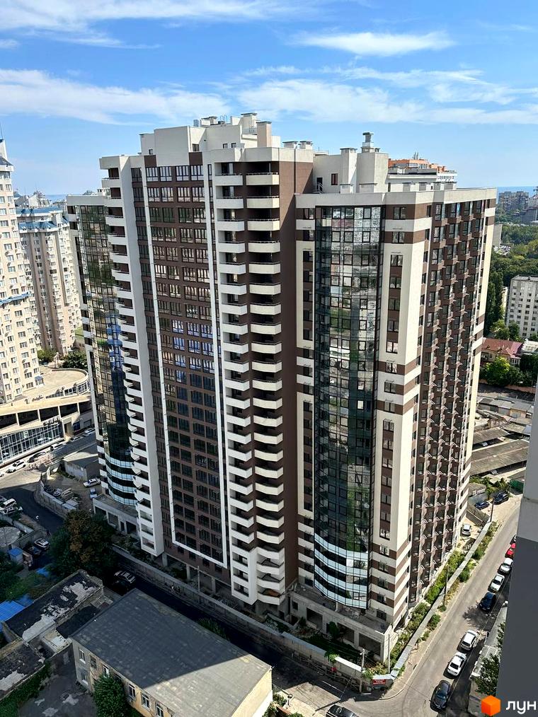 Зовнішній вигляд житлового комплексу Удобний з коричнево-білою фасадною обробкою та скляними балконами. Навколо комплексу розташована дорога з припаркованими автомобілями.