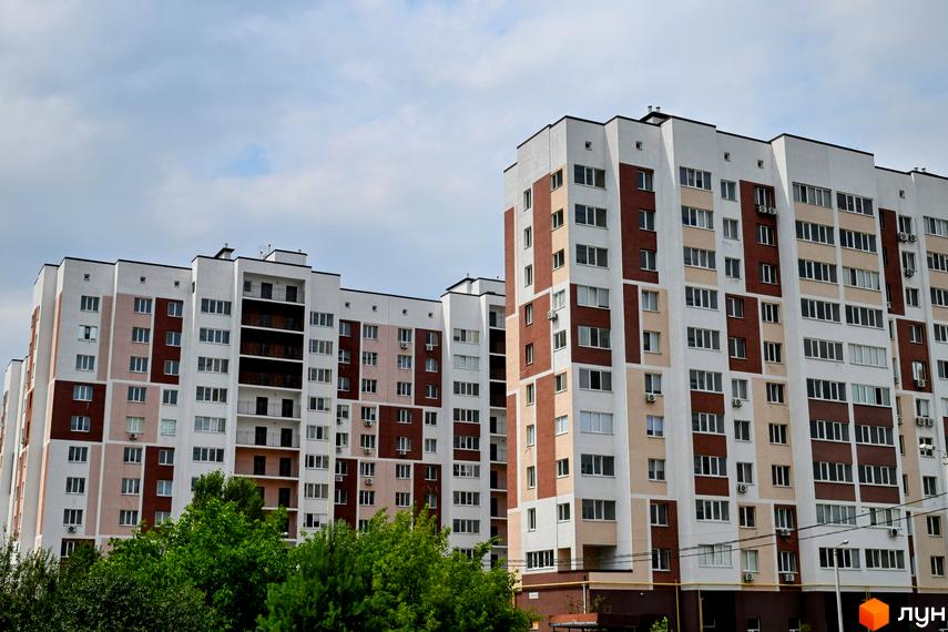 Фасад житлового комплексу Сади Вишневі з білими та коричневими елементами, багато вікон та балконів. Навколо зелені дерева.