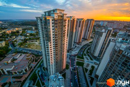 Високі житлові будинки Житлового комплексу Новопечерські Липки з світлими фасадами та великими вікнами. Навколо будинків видно дороги, парковки з автомобілями та зелені зони.