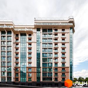 Житловий комплекс HydroPark DeLuxe: багатоповерховий будинок світлого кольору з великими вікнами та балконами. На передньому плані видно парковку, а на задньому – зелені насадження.