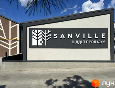 Вивіска ЖК Sanville з логотипом у вигляді дерев. На фоні будівля з білими стінами та темним обрамленням, бруківка перед будівлею.