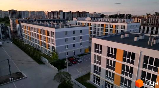 Зовнішній вигляд новобудови ЖК Sanville Park з сірими та жовтими фасадами, вікнами та парковкою. Навколо комплексу зелені насадження та сіра огорожа.