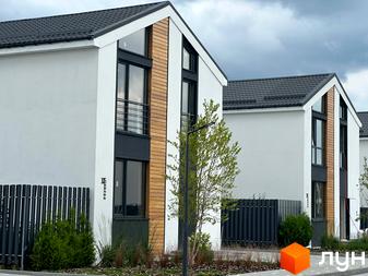 Котеджне містечко Gardd House Eco Village. Фасад будинку з білою штукатуркою, дерев'яними вставками та темними вікнами. Поруч паркан та зелені насадження.