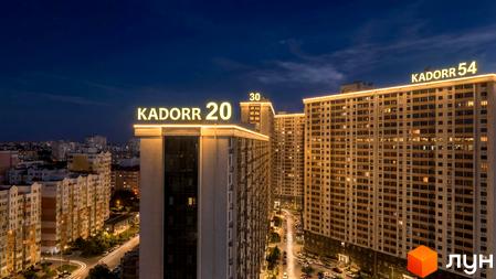 Житловий комплекс KADORR 20 з сучасними фасадами, освітленими вночі. Прибудинкова територія з парковими зонами та автомобільними стоянками.