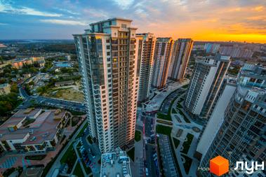 Високі житлові будинки Житлового комплексу Новопечерські Липки з світлими фасадами та великими вікнами. Навколо будинків видно дороги, парковки з автомобілями та зелені зони.