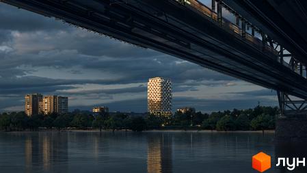 Висотна будівля житлового комплексу Kub29 з великою кількістю вікон, розташована біля річки. Навколо будинку зелені дерева та спокійна вода.