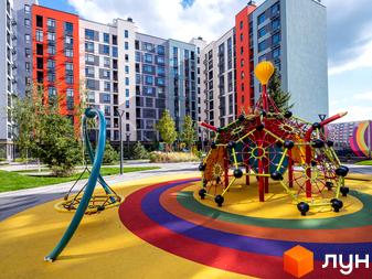 Житловий комплекс Оптимісто з яскравим дитячим майданчиком та кольоровим покриттям. Будинки мають різнокольорові фасади та багато вікон.