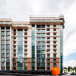 Житловий комплекс HydroPark DeLuxe: багатоповерховий будинок світлого кольору з великими вікнами та балконами. На передньому плані видно парковку, а на задньому – зелені насадження.