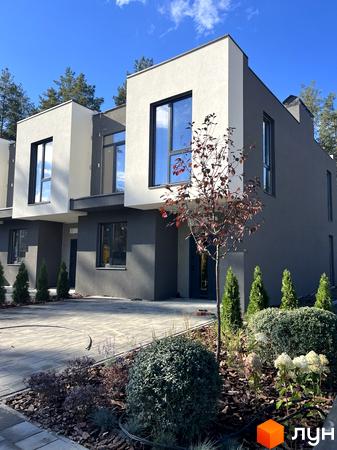 Будинок з сірим та білим фасадом та великими вікнами. Прибудинкова територія з зеленими насадженнями та бруківкою.