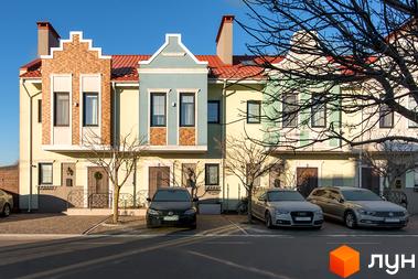 Котеджне містечко КМ Balaton Village. Фасади будинків у світлих тонах з червоною черепицею, перед будинками припарковані автомобілі та дерева без листя.