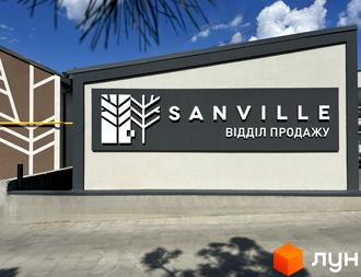 Вивіска ЖК Sanville з логотипом у вигляді дерев. На фоні будівля з білими стінами та темним обрамленням, бруківка перед будівлею.