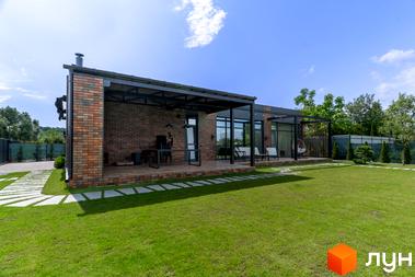 Котеджне містечко КМ Озерний: одноповерховий будинок з цегляним фасадом та терасою, доглянутий зелений двір з доріжками та рослинами.